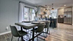A modern kitchen with a dining area, featuring a wooden table, bar stools, and stainless steel appliances. - Decatur Rocksteady Message for Summer Special (Decatur)