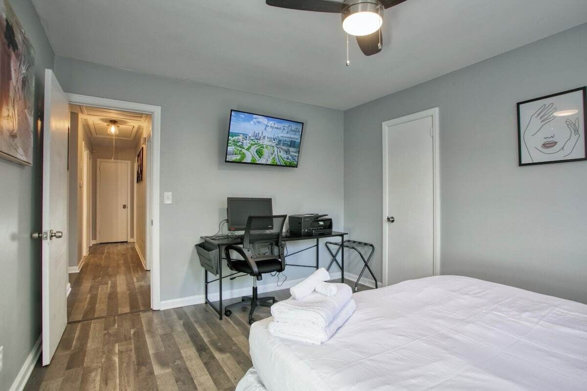 A hotel room with a bed, a desk with a computer, a chair, a television on the wall, and a door leading to another room.