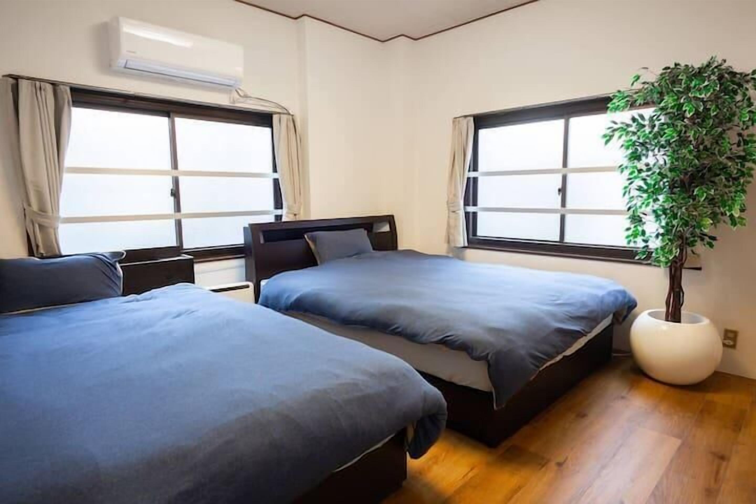 A bedroom with two beds, large windows, a potted plant, and an air conditioning unit.