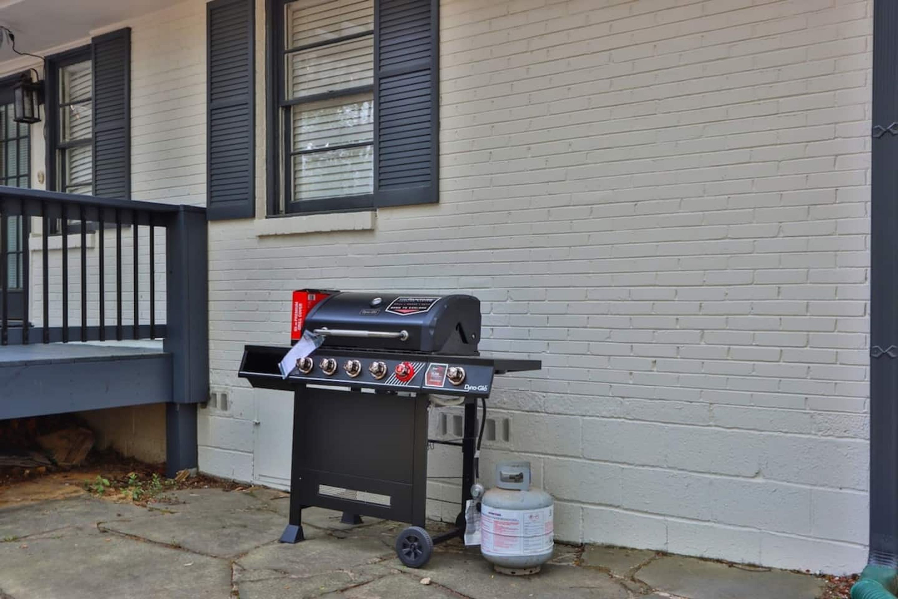 A portable grill with a gas tank and a control panel, placed on a concrete surface next to a white brick building with black shutters.