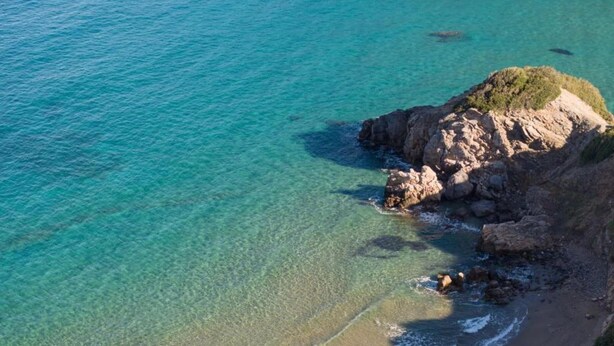 Vista aérea de una zona costera con aguas turquesas cristalinas, acantilados rocosos y una pequeña isla con vegetación.
