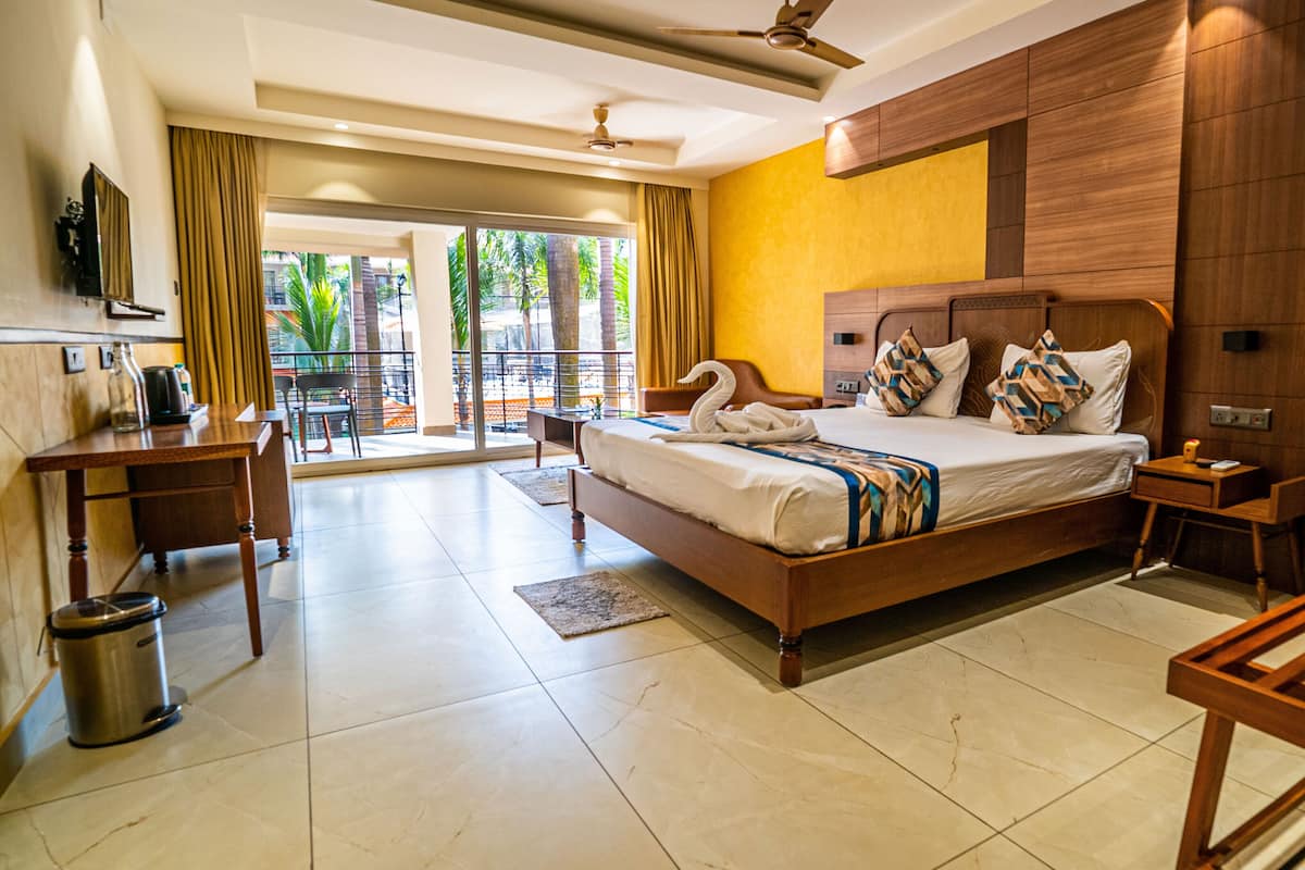 Habitación de hotel con una cama grande, cabecera de madera, escritorio con silla, televisor y balcón con vista a palmeras.