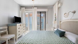 Une chambre à coucher avec un lit, un téléviseur posé sur une commode, une chaise, une autre commode et une fenêtre avec des rideaux.