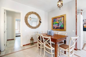 Un comedor con una mesa de madera, sillas blancas, un espejo decorativo y un cuadro colorido en la pared.