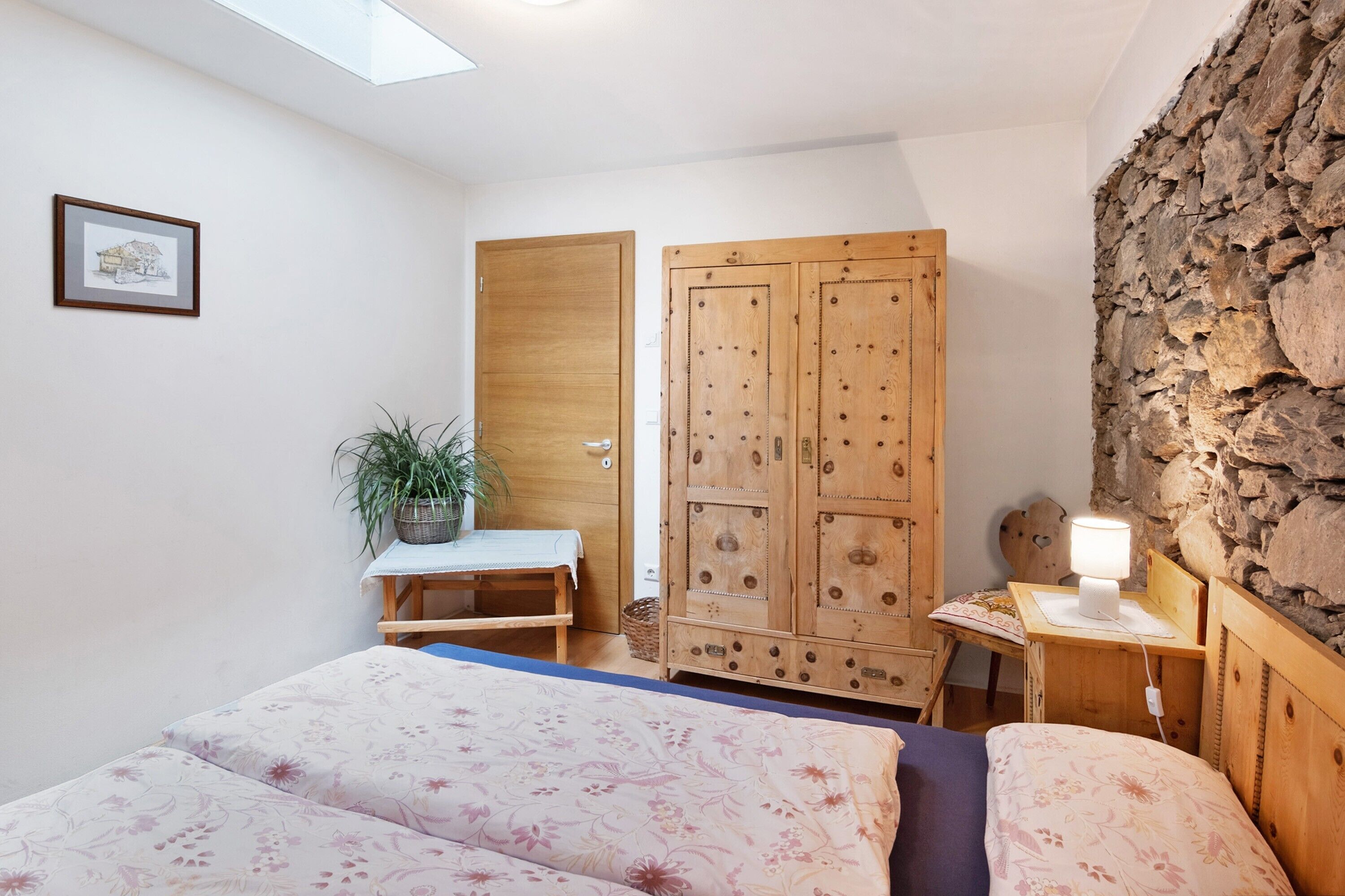 Un dormitorio con dos camas, un armario de madera, un escritorio con lámpara, una planta en maceta y una pared de piedra.