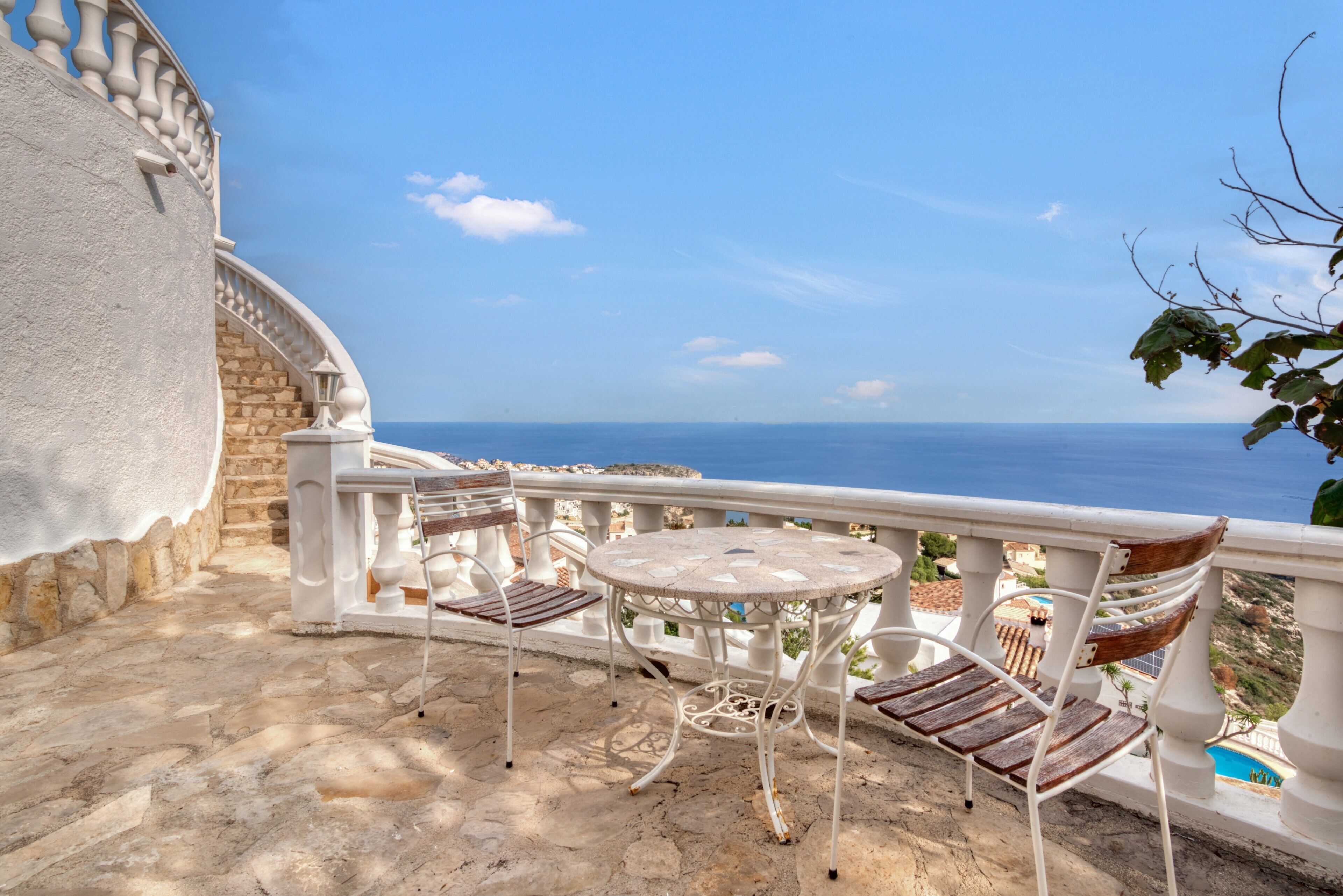 A stone staircase leading to a balcony with a round table and two chairs, overlooking a coastal view.