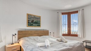 A bedroom with a wooden headboard, a bed with a patterned bedspread, two rolled towels, a nightstand with a lamp, a painting of a seascape, a window with curtains, and a view of the sea. - Villa 'Costa Blanca' with Sea View, Wi-Fi and Air Conditioning (Benitachell)