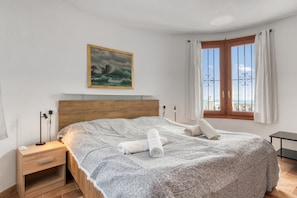 Ein Schlafzimmer mit hölzernem Kopfteil, einem Bett mit gemustertem Überwurf, zwei aufgerollten Handtüchern, einem Nachttisch mit Lampe, einem Seestück als Gemälde, einem Fenster mit Vorhängen und Blick auf das Meer.