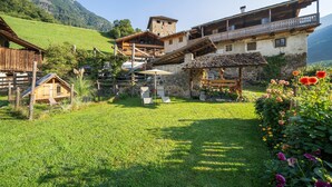 A traditional alpine wooden house with a stone tower, a wooden balcony, and a garden with flowers.