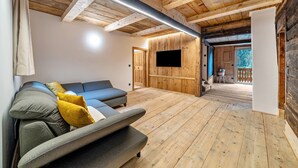 A room with wooden flooring, a sofa with cushions, a flat-screen TV mounted on a wooden wall, and a door leading to an outdoor area.