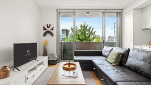Una sala moderna con televisor de pantalla plana, una mesa de centro de madera, un sofá gris y un ventanal corredizo que da a un balcón con vistas a la ciudad.
