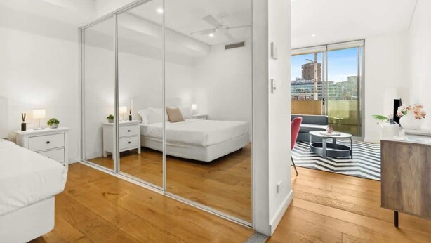 A modern bedroom with a glass wardrobe, a bed, bedside tables, and a view of the city through large windows.