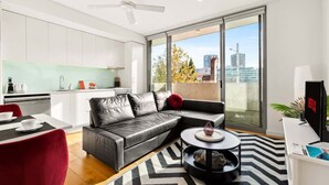 A modern living room with a black leather sofa, a dining table with red chairs, a black coffee table, and a large window offering a city view.