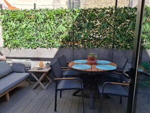 A patio area with a round table, blue chairs, a grey sofa, and a green leafy wall.