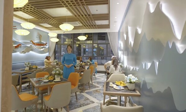 A restaurant with tables and chairs, a woman in a blue dress, and a wall with a mountain design. - The Moon Boutique Hotel Cat Ba (Hai Phong)