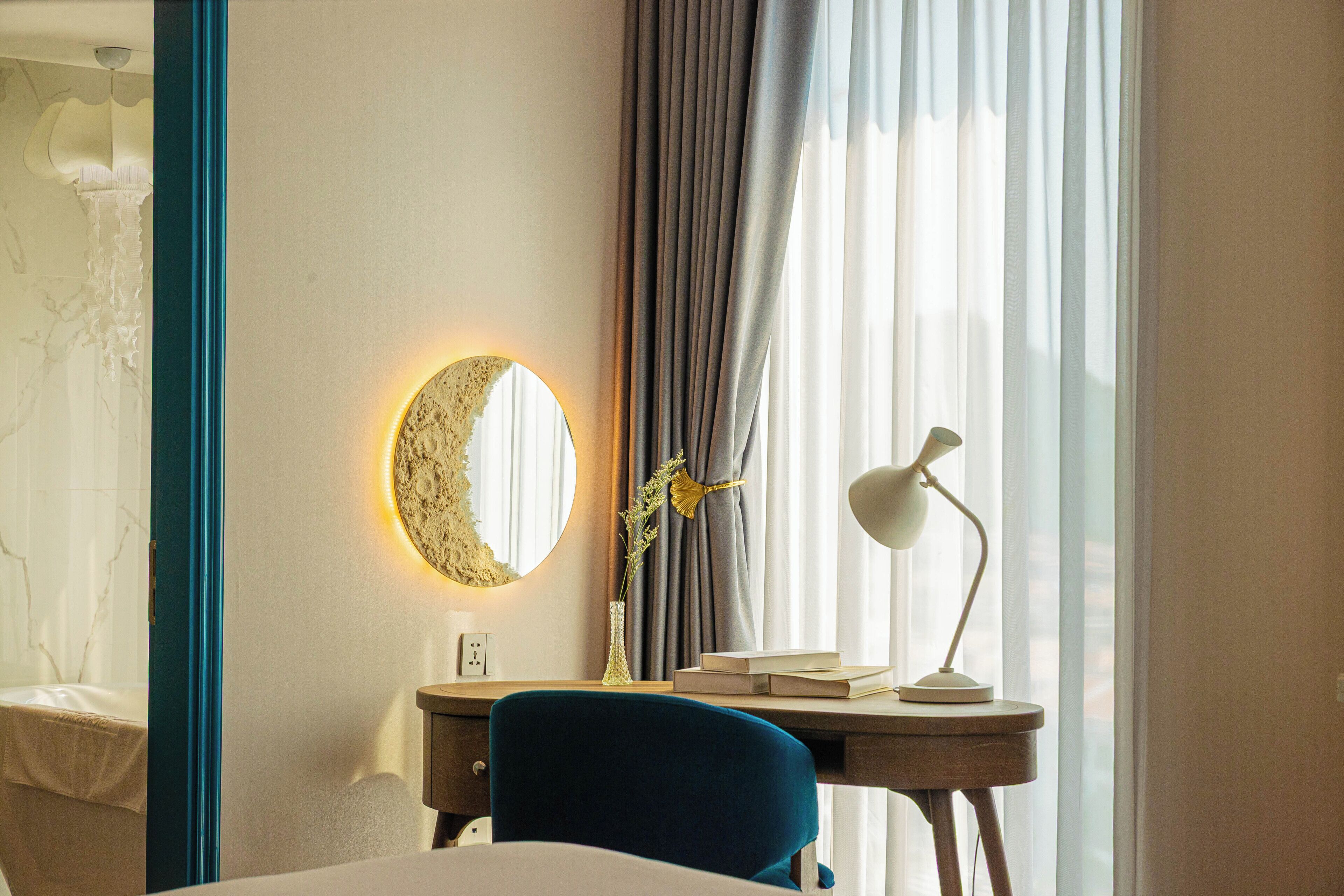 A hotel room with a bed, a desk with a lamp, a mirror, and a window with curtains.