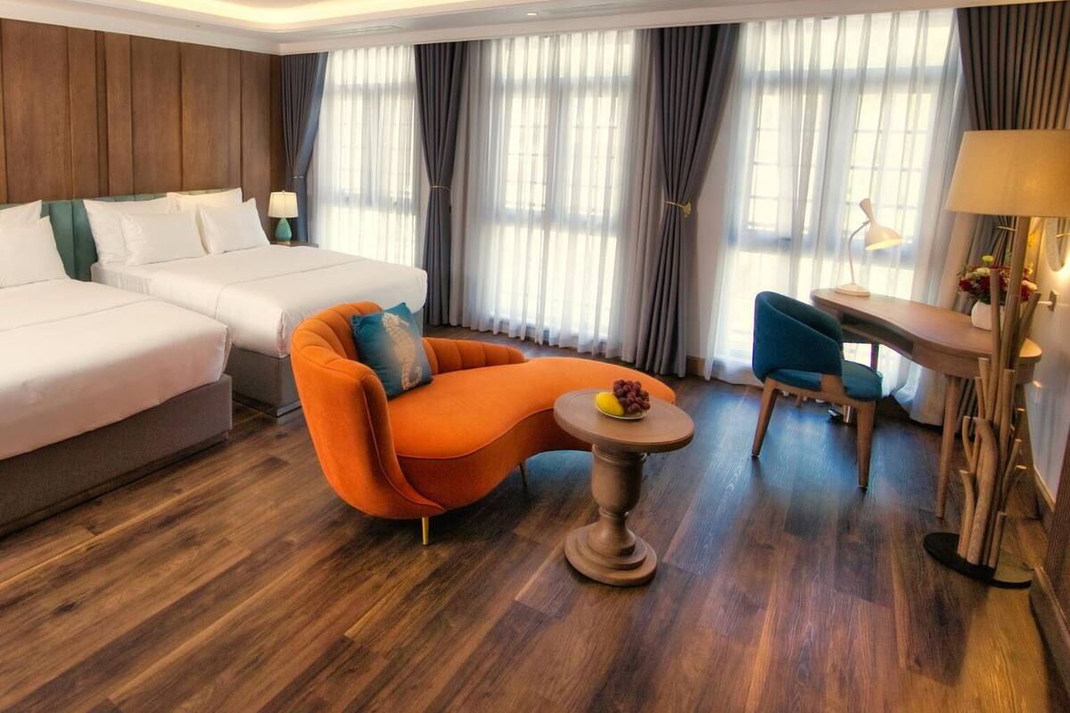 A hotel room with two beds, a modern orange armchair, a small round table, and a desk with a chair.