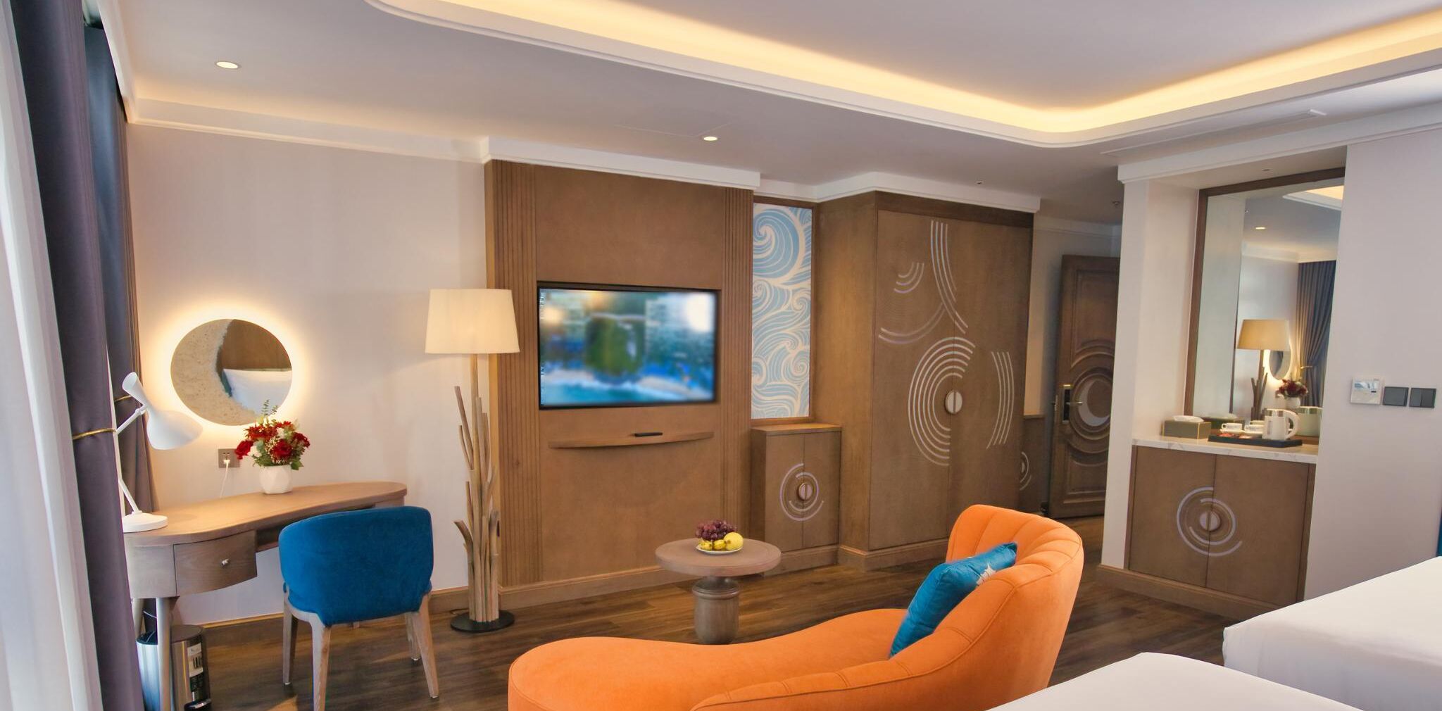 A modern hotel room with a TV, a desk with a lamp, a blue chair, an orange sofa, and a small table with fruit.