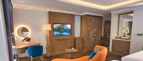 A modern hotel room with a TV, a desk with a lamp, a blue chair, an orange sofa, and a small table with fruit.
