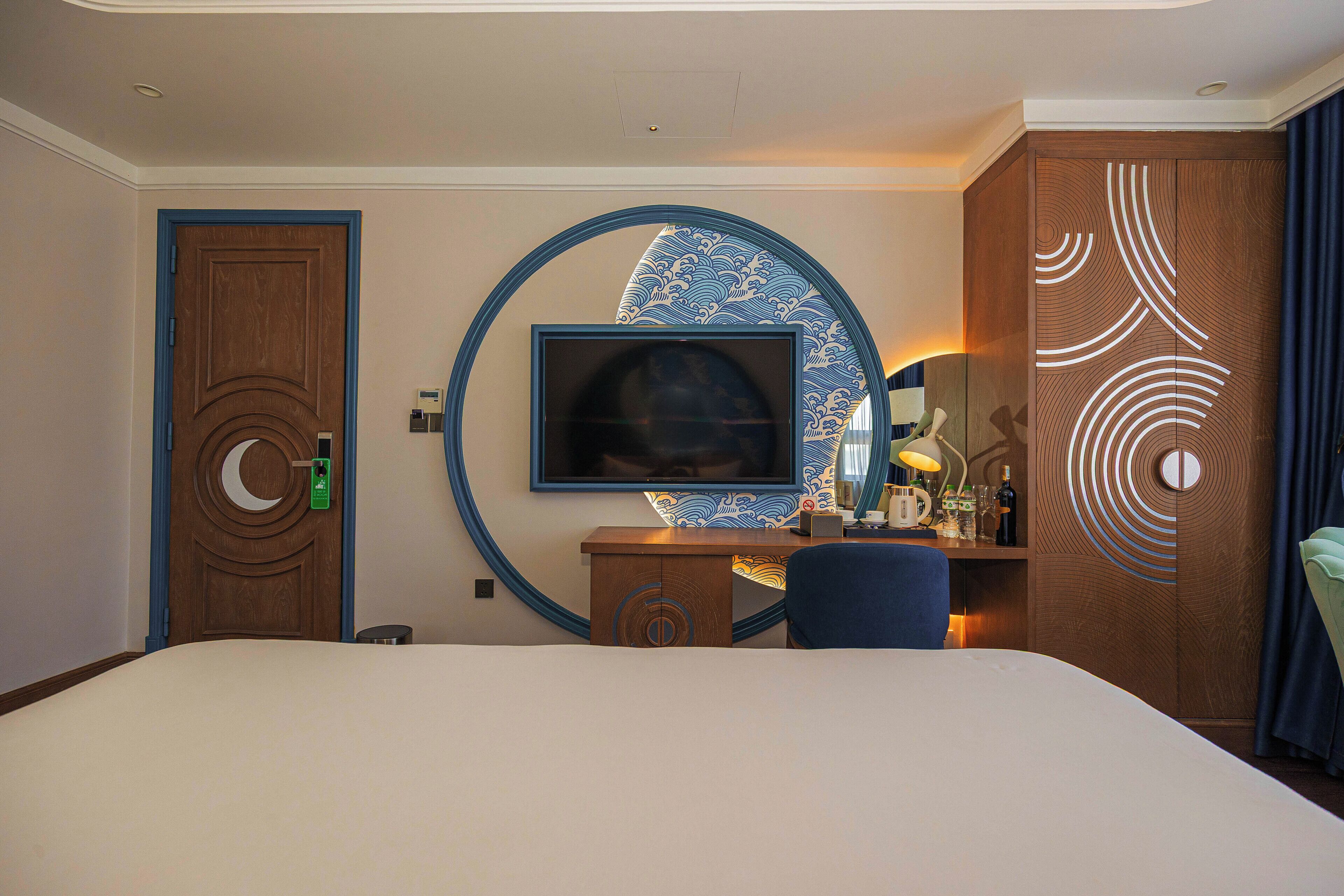 A hotel room with a large bed, a television mounted on the wall, a desk with a chair, and a decorative wall panel.