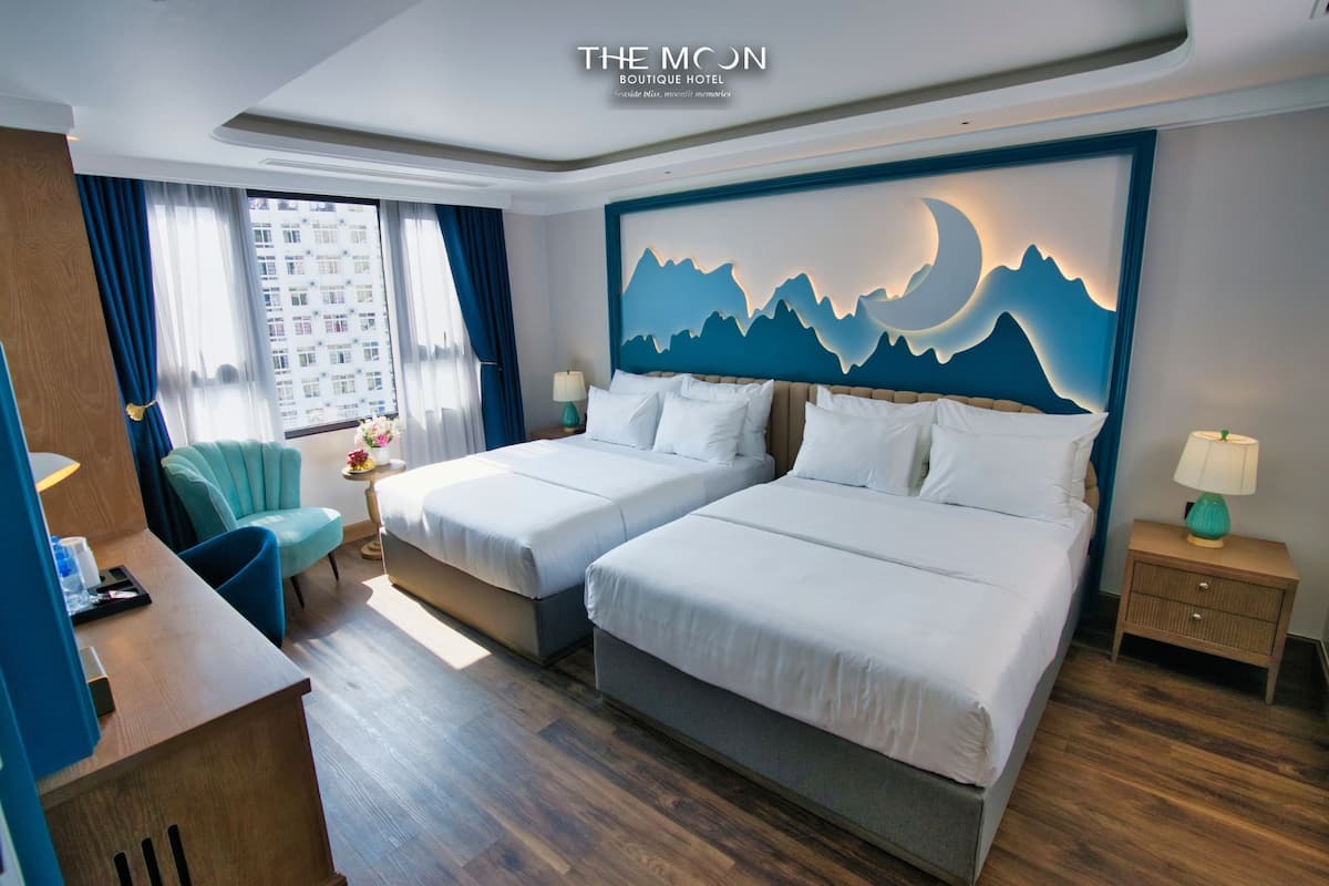 A hotel room with two beds, a desk, a chair, a nightstand, and a wall art depicting mountains and a moon.