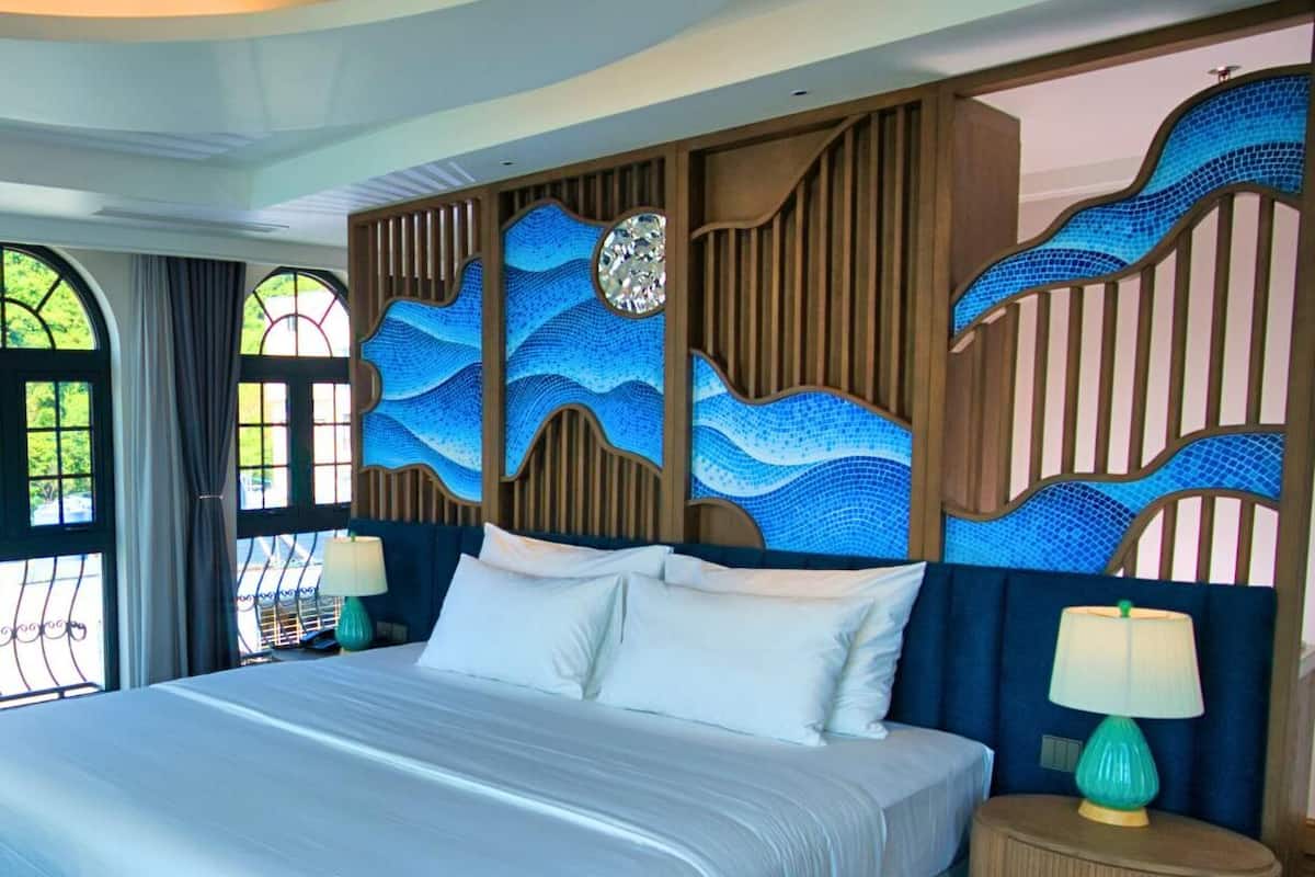 A large bed with a wooden headboard, a nightstand with a lamp, and a view of greenery outside.