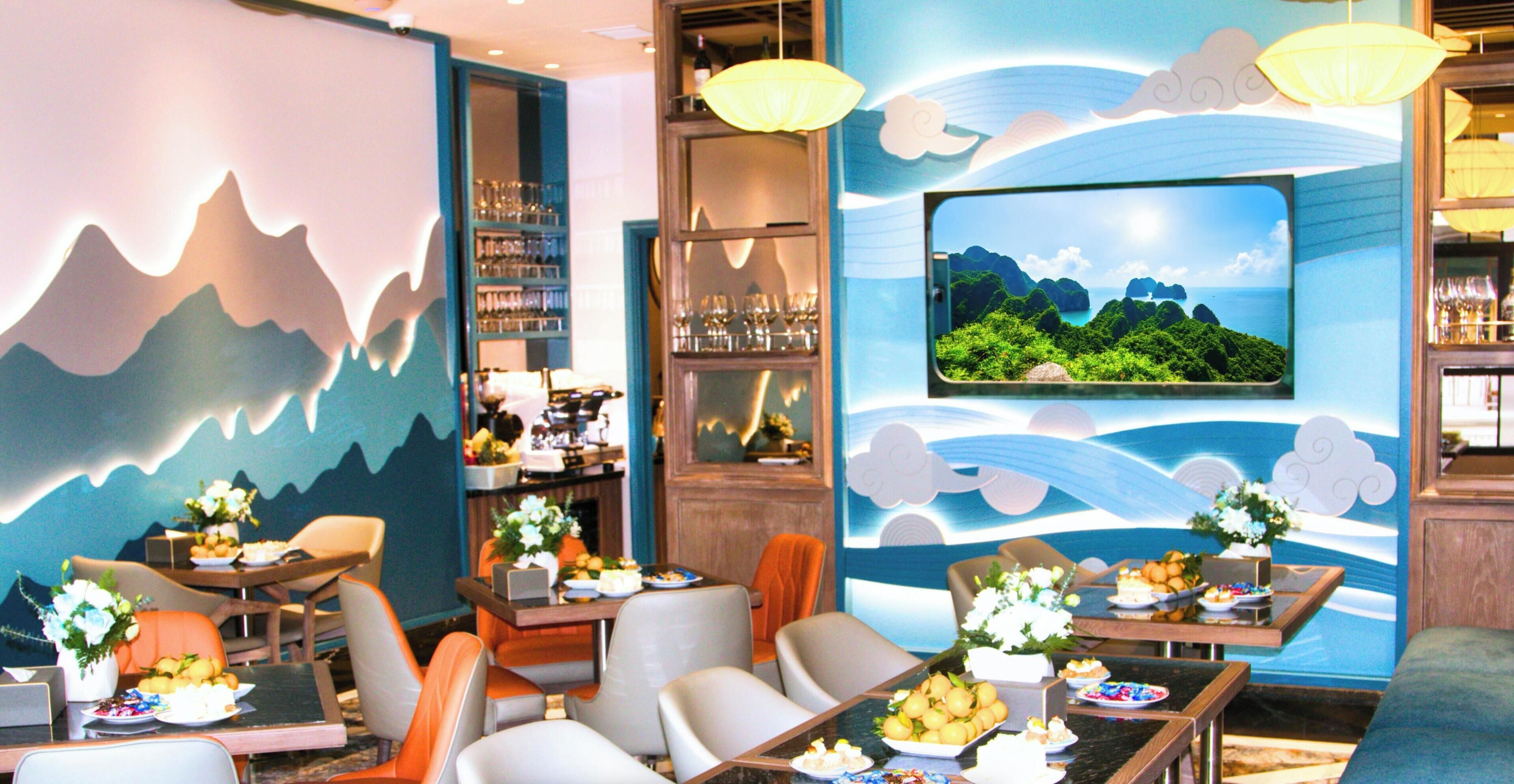 Un restaurante con un mural temático de montaña, una pantalla de televisión que muestra un paisaje tropical y diversos platos sobre las mesas.