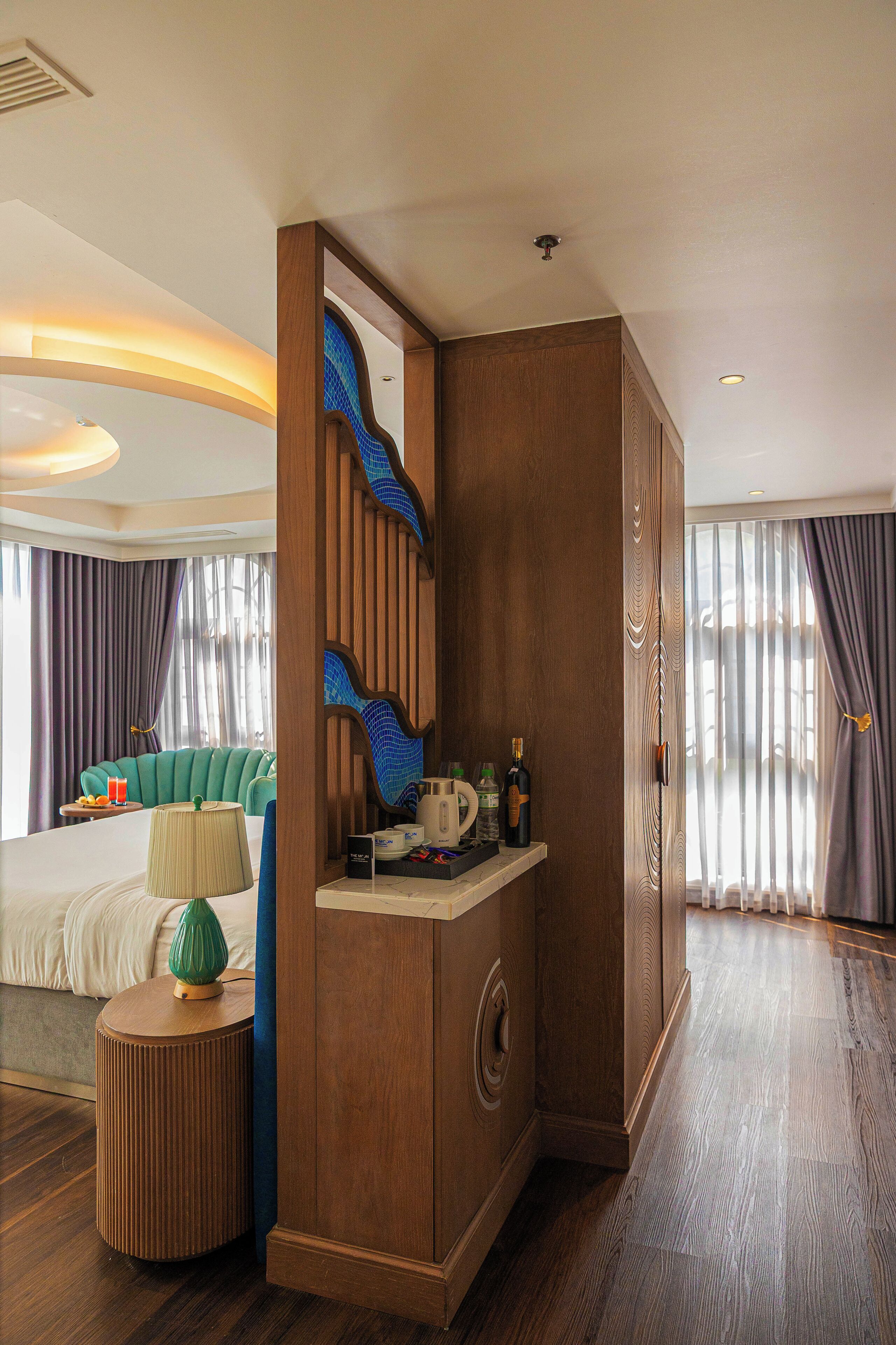 A hotel room with a wooden partition, a bed with a green headboard, a small round table with a lamp, and a minibar with various items.