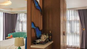 A hotel room with a wooden partition, a bed with a green headboard, a small round table with a lamp, and a minibar with various items.