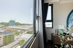 Una habitación con vista a un paisaje urbano, una ventana con cortinas, una mesita de noche con una lámpara y un plato decorativo.