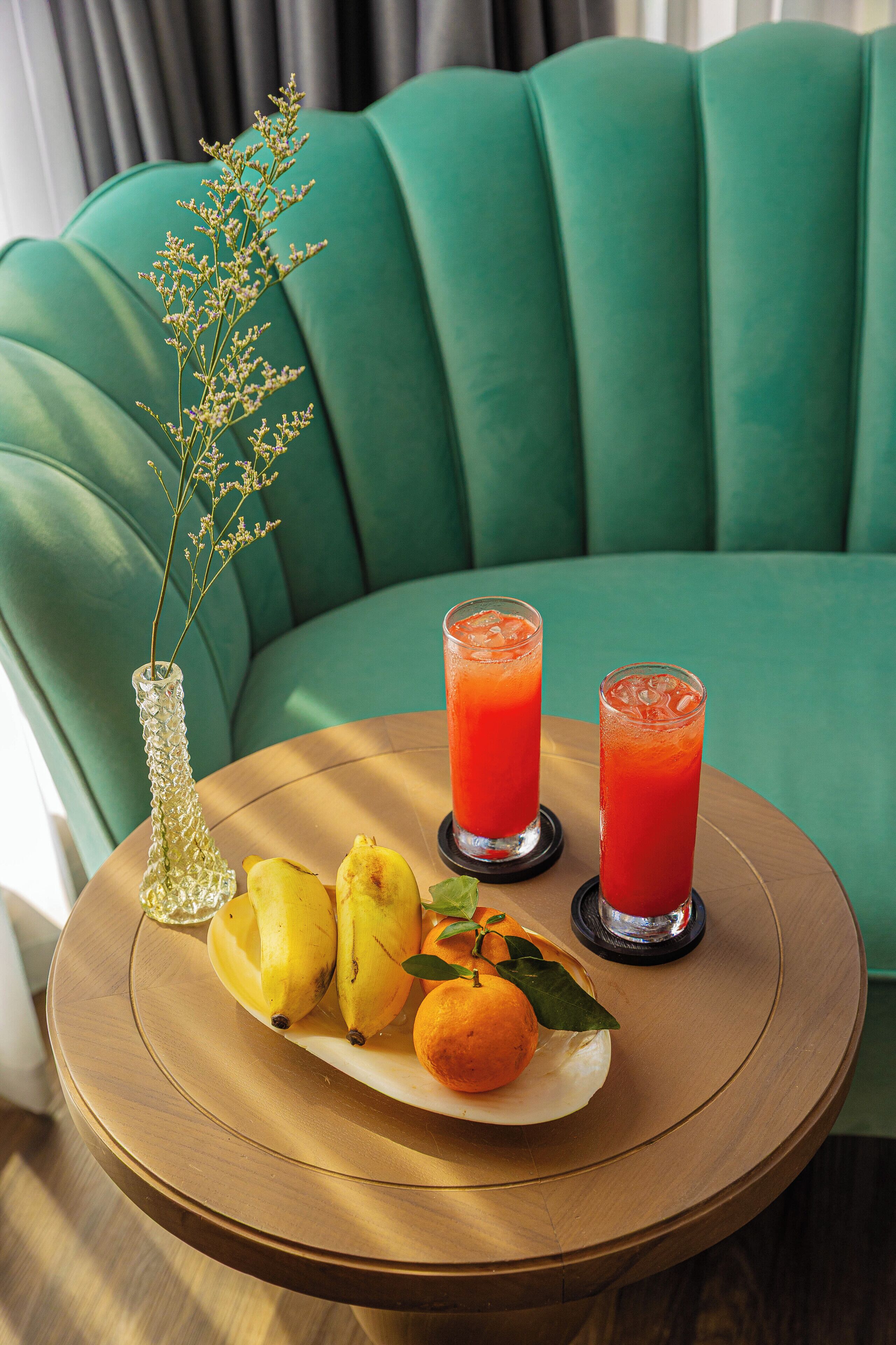A round wooden table with a plate of fruit, two glasses of red drink, and a decorative vase with a plant.