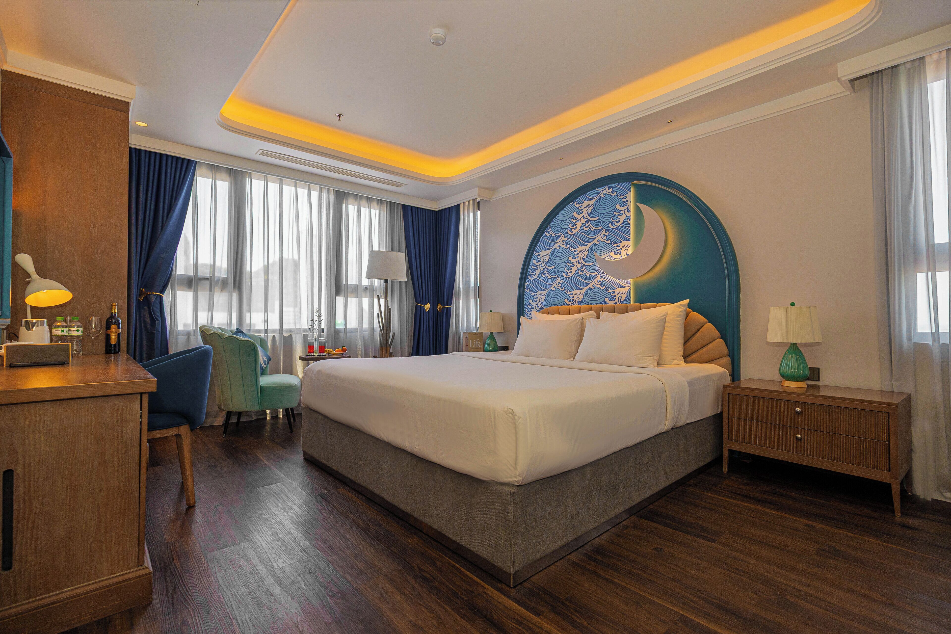 A hotel room with a large bed, a desk with a lamp, a chair, and a nightstand with a lamp.