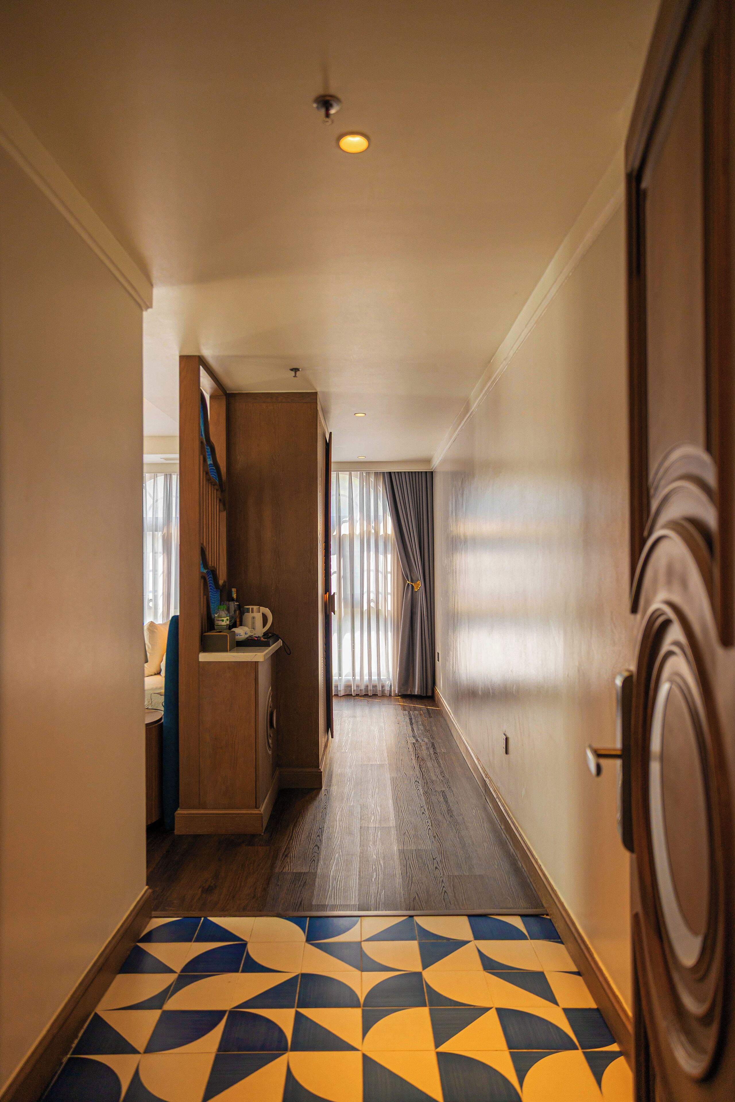 Een hotelhal met een vloer met patroon, een houten deur en een doorkijkje naar een kamer met raam en gordijnen.