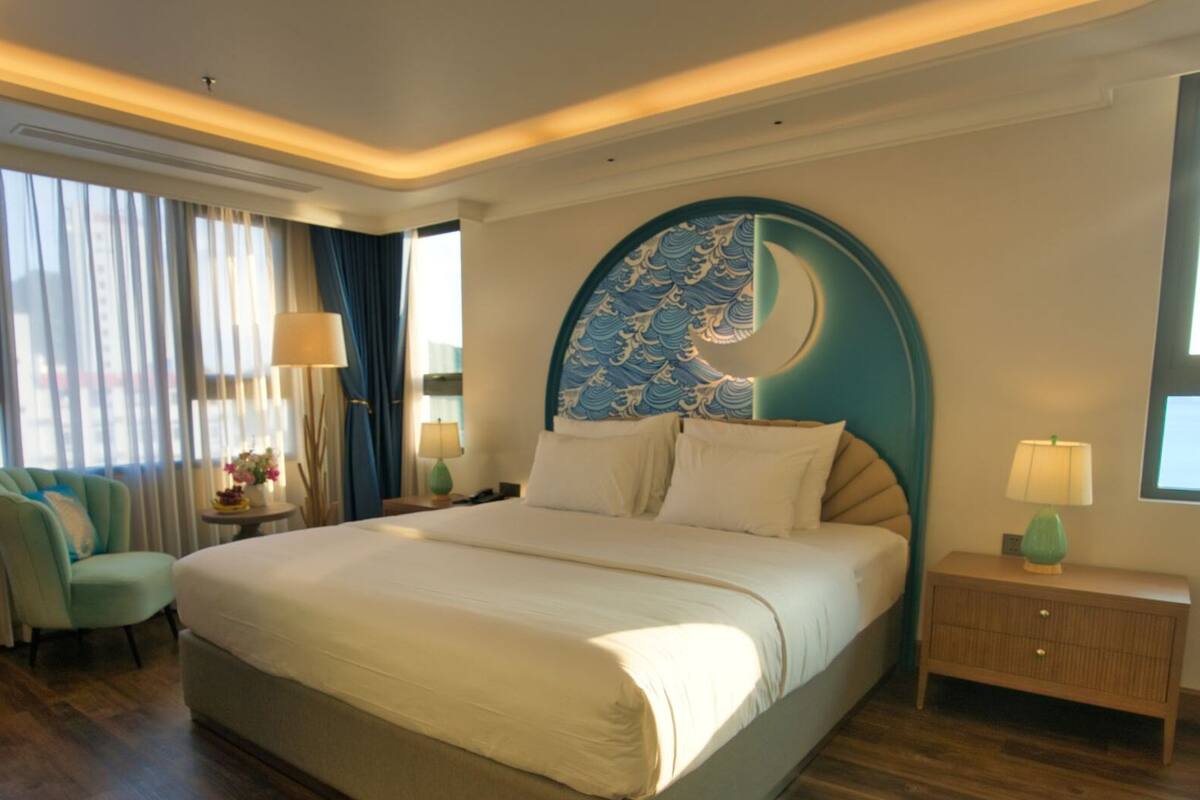 A hotel room with a large bed, a curved headboard with a wave pattern, a desk, a chair, a nightstand with a lamp, and a window with curtains.