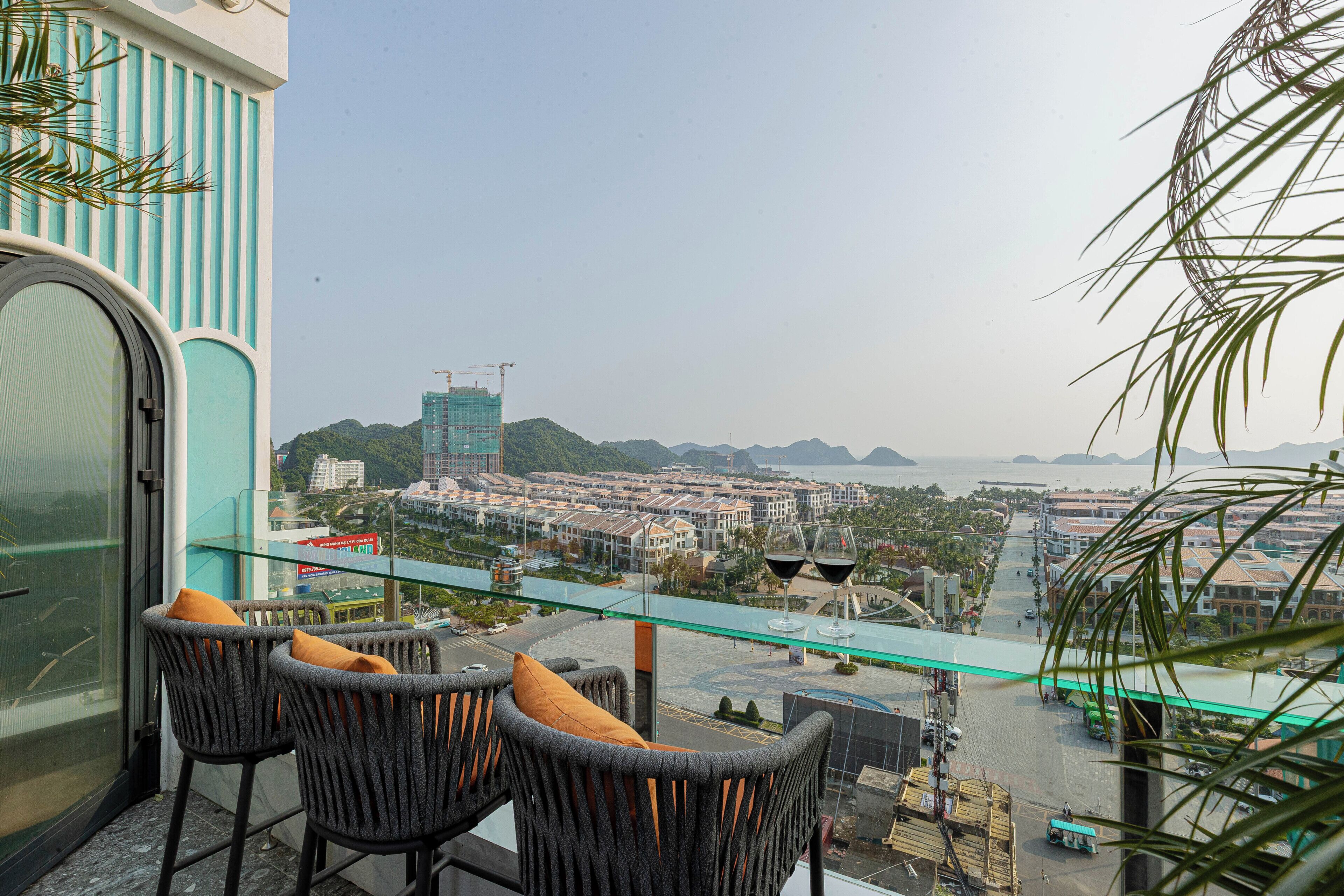 A balcony with wicker chairs and a glass railing overlooking a cityscape with buildings and a bridge.