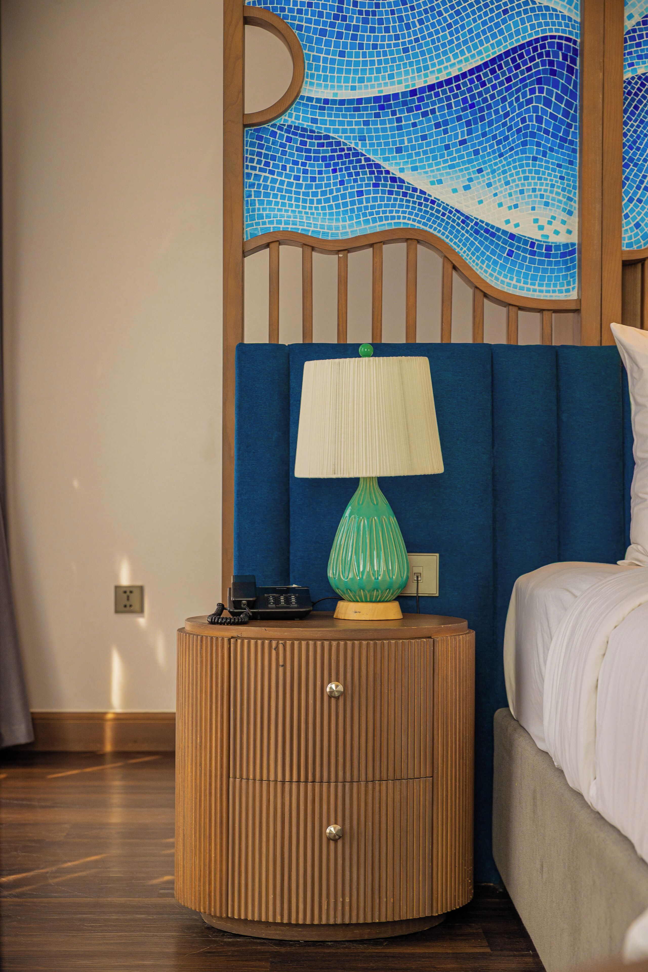 Une chambre d’hôtel avec une table de chevet en bois, une lampe verte, un téléphone et un lit avec une literie blanche.