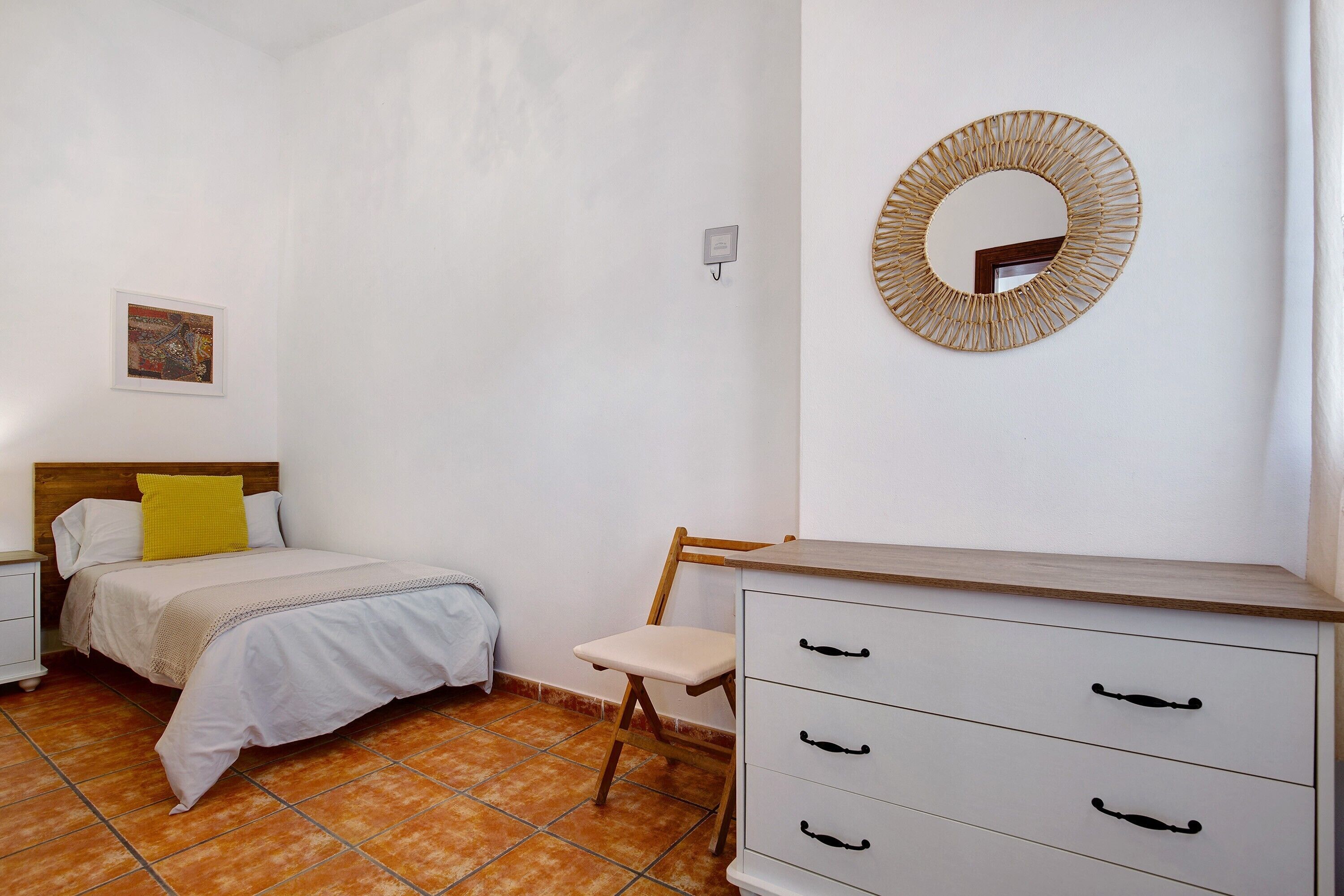 Un dormitorio con cama, sillón, cómoda, espejo en la pared y un cuadro.