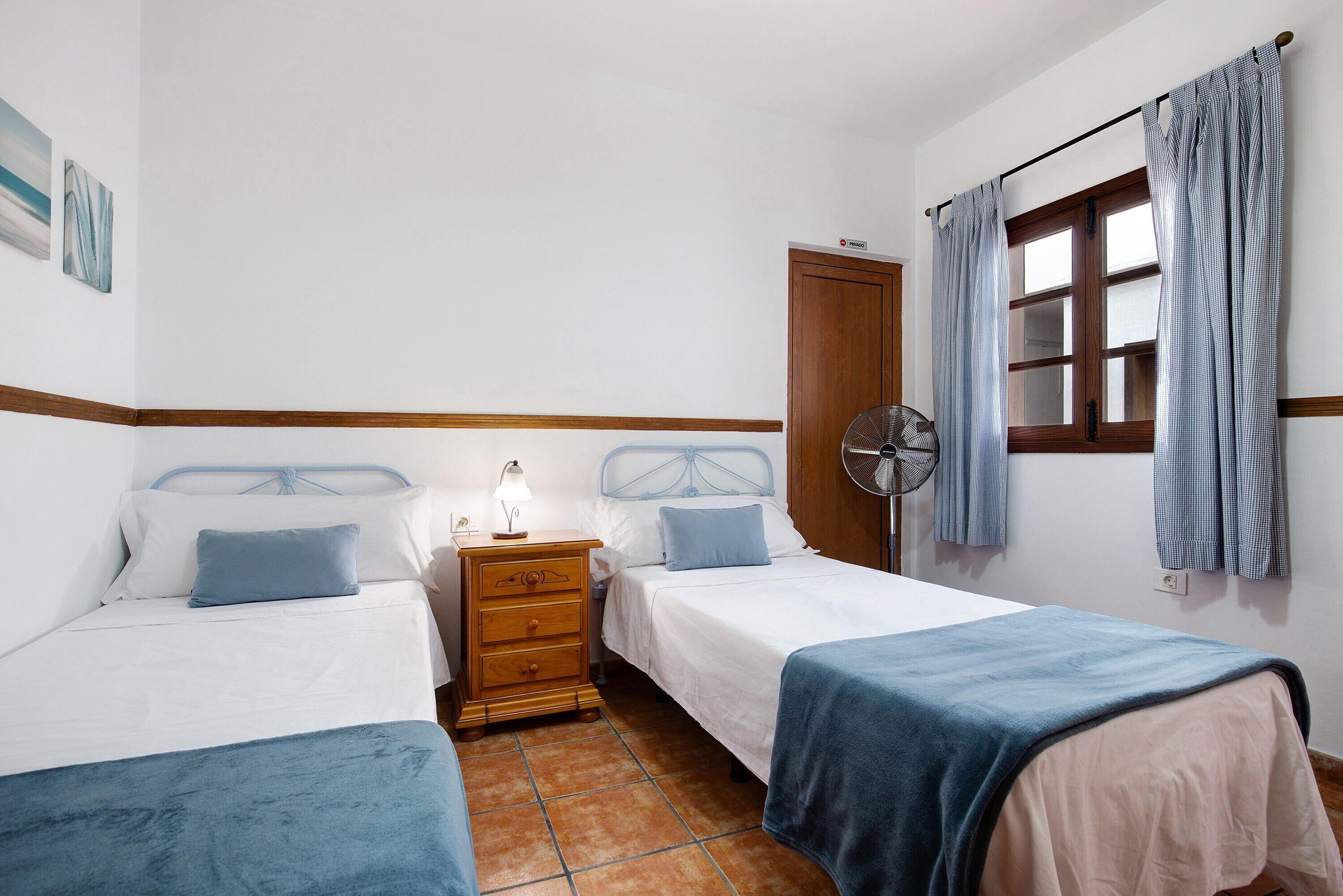 A bedroom with two beds, a nightstand with a lamp, a fan, and a window with curtains.
