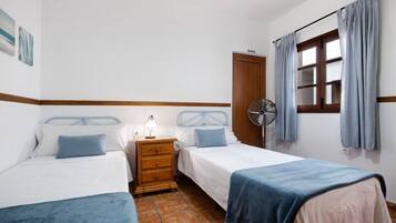 A bedroom with two beds, a nightstand with a lamp, a fan, and a window with curtains.