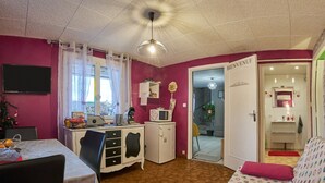 Ein Zimmer mit einer Küchenzeile, einem Essbereich, einem Bett mit gemustertem Bezug, einer weißen Tür mit der Aufschrift 'BIENVENUE' und einem Deckenventilator.