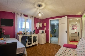 A room with a kitchenette, a dining area, a bed with a patterned cover, a white door with 'BIENVENUE' sign, and a ceiling fan.