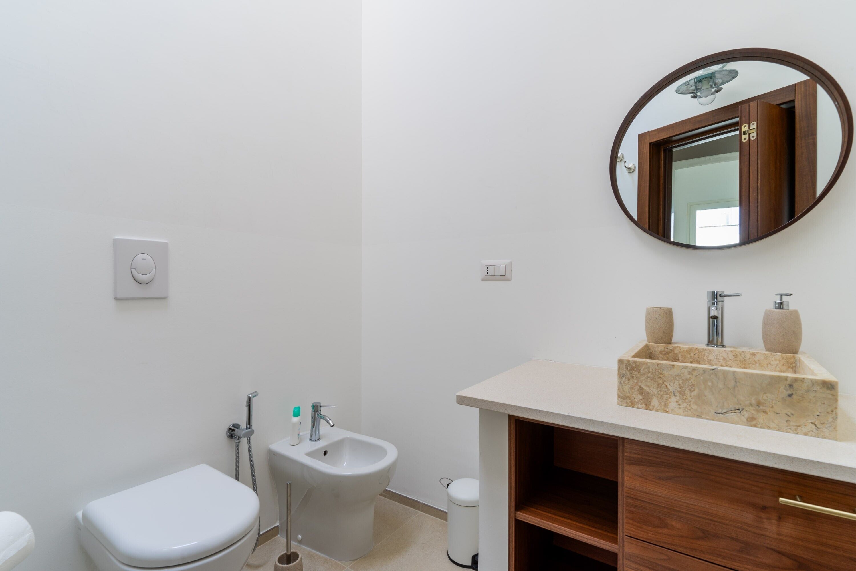 Een badkamer met toilet, bidet, wastafel, spiegel en een kleine prullenbak.
