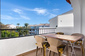 Un balcón con una mesa y sillas, una pared blanca y vistas a edificios y árboles.