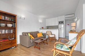 Un espacio habitable compacto con sofá, una mesa de comedor, una silla de mimbre, una estantería y una kitchenette.