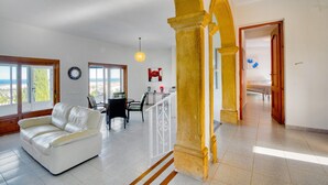 A spacious living area with a white leather sofa, a dining table with chairs, a kitchen area, and a view through large windows. - Villa 'Alta' with Private Pool, Wi-Fi and Air Conditioning (Dénia)