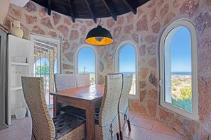 A stone-walled room with a wooden table and wicker chairs, large arched windows, and a view of the landscape.