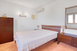 A bedroom with a bed, wooden dresser, bedside lamps, an air conditioner, and a wall-mounted light fixture.