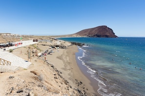 A beach with a rocky shoreline, a clear blue sea, and a prominent hill in the background.