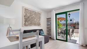 Una habitación con una mesa blanca, un sofá gris, un cuadro abstracto grande y un ventanal corredero que da a un patio exterior con mesa y sillas.