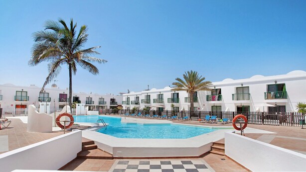 Zona de piscina con suelo a cuadros, palmeras y un edificio blanco con balcones.
